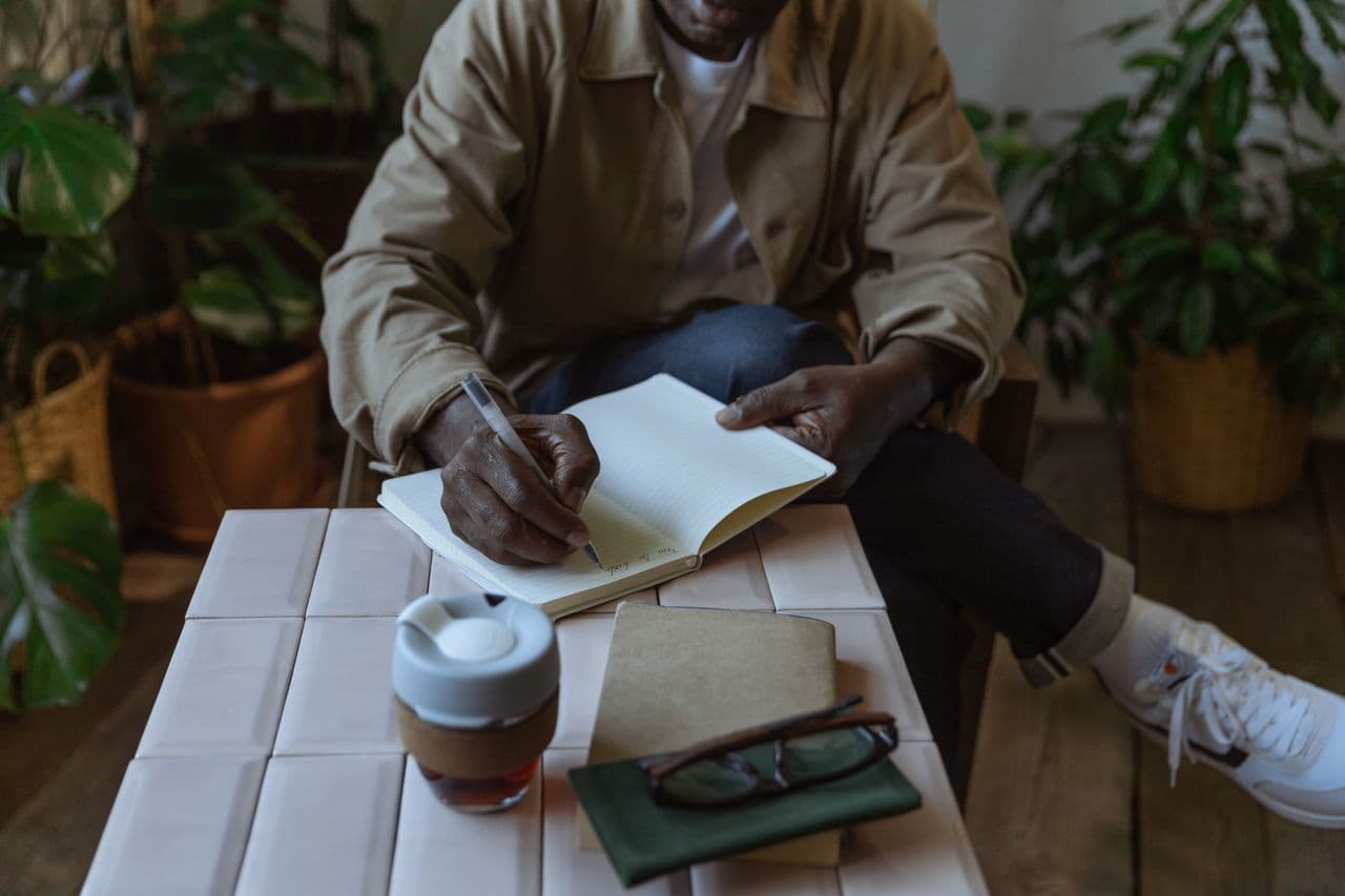 Someone writing in a lined notebook at a tiled table, a coffee cup and reading glasses beside them, houseplants behind